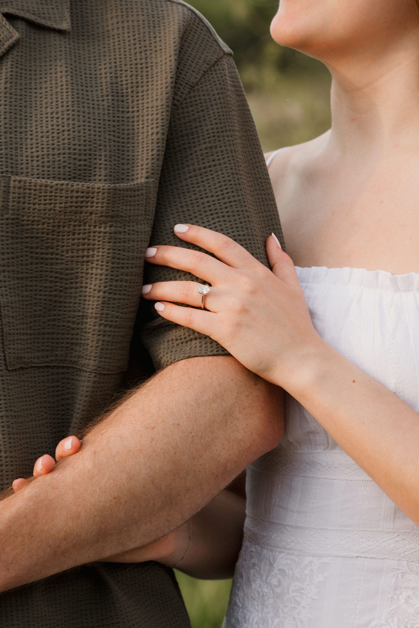 Close-up of a bride in white dress holding groom's arm, showcasing her diamond engagement ring.