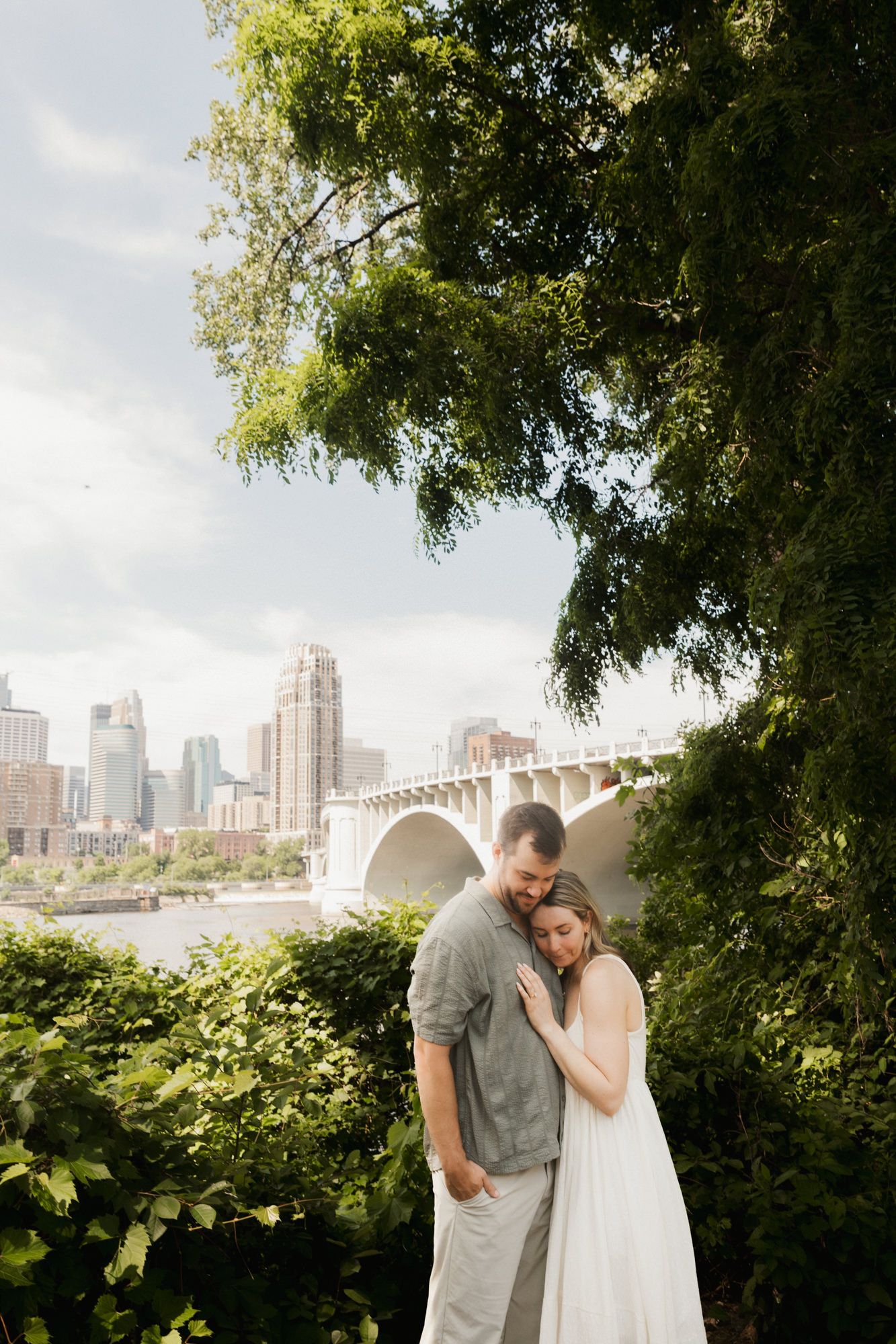 Couple embracing outdoors with lush greenery, a bridge, and city skyline in the background on a sunny day.