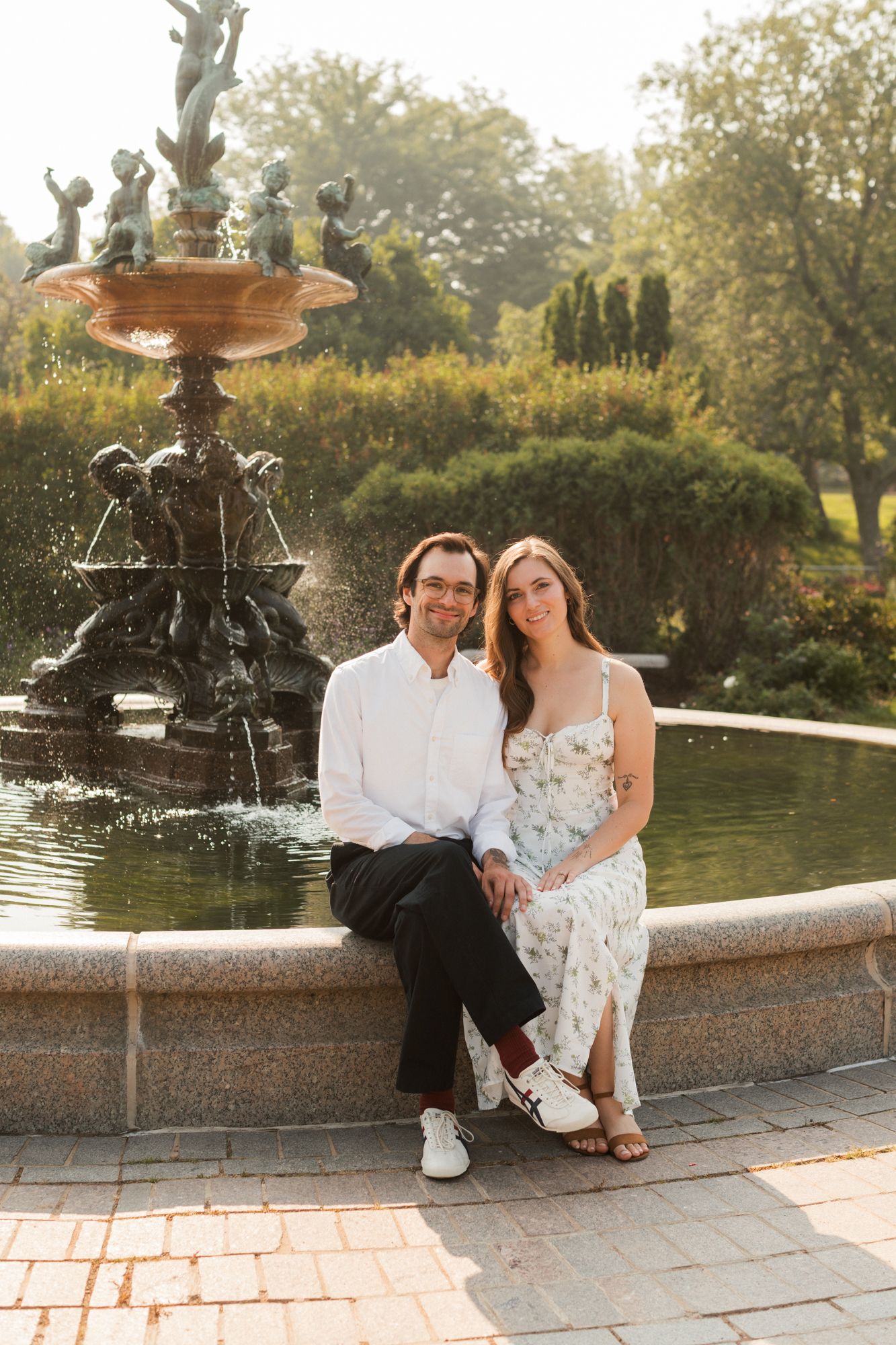 Couple sitting together by an ornate fountain in a sunlit park, man in white shirt and woman in floral dress smiling.