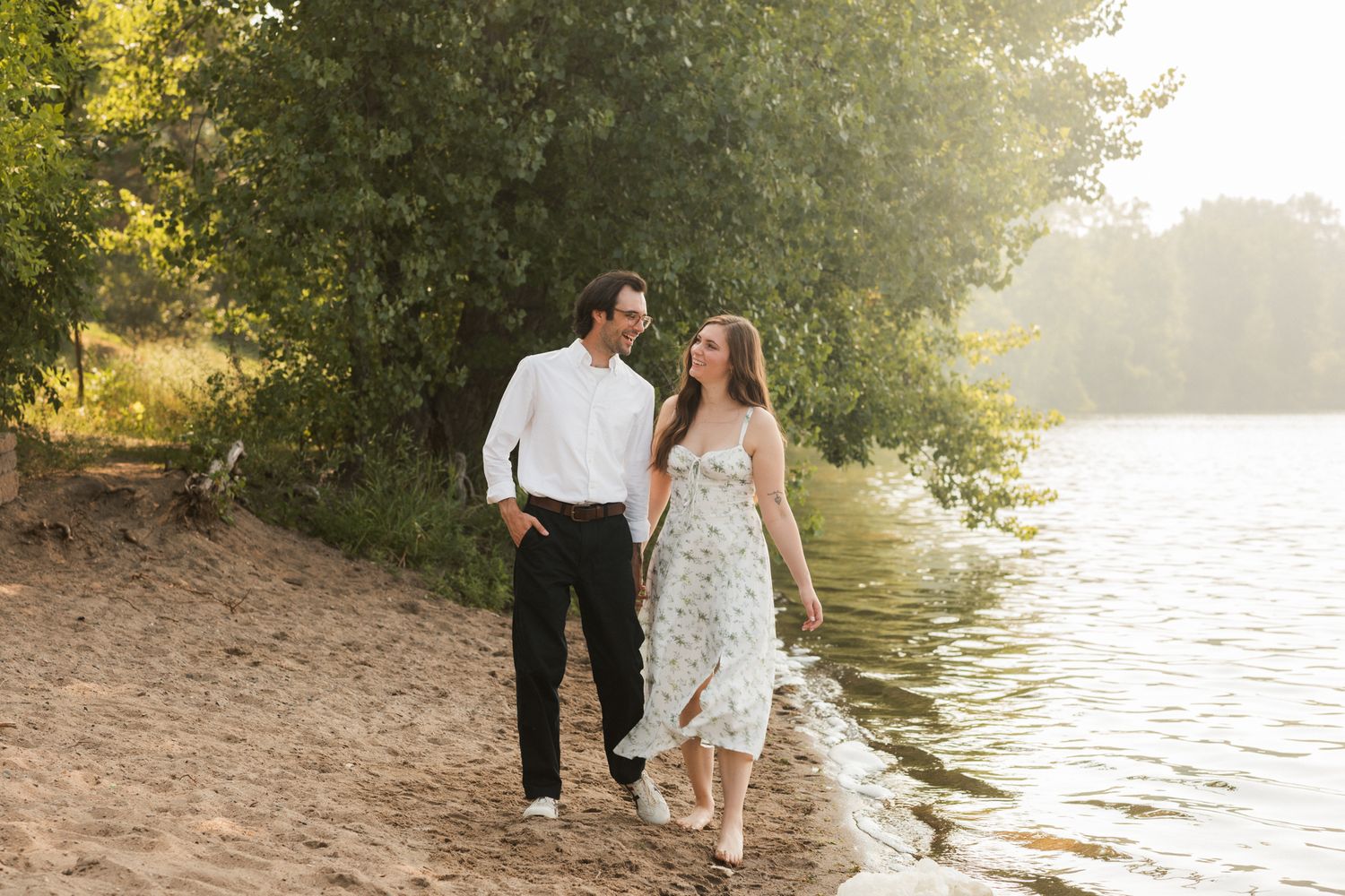 Couple walking hand in hand along a sandy lakeside beach surrounded by lush green trees in golden hour light.