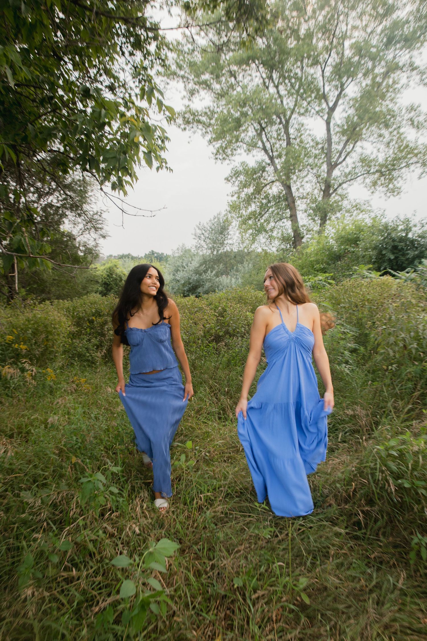 Two women in matching blue maxi dresses walk through a lush green outdoor setting, smiling at each other.