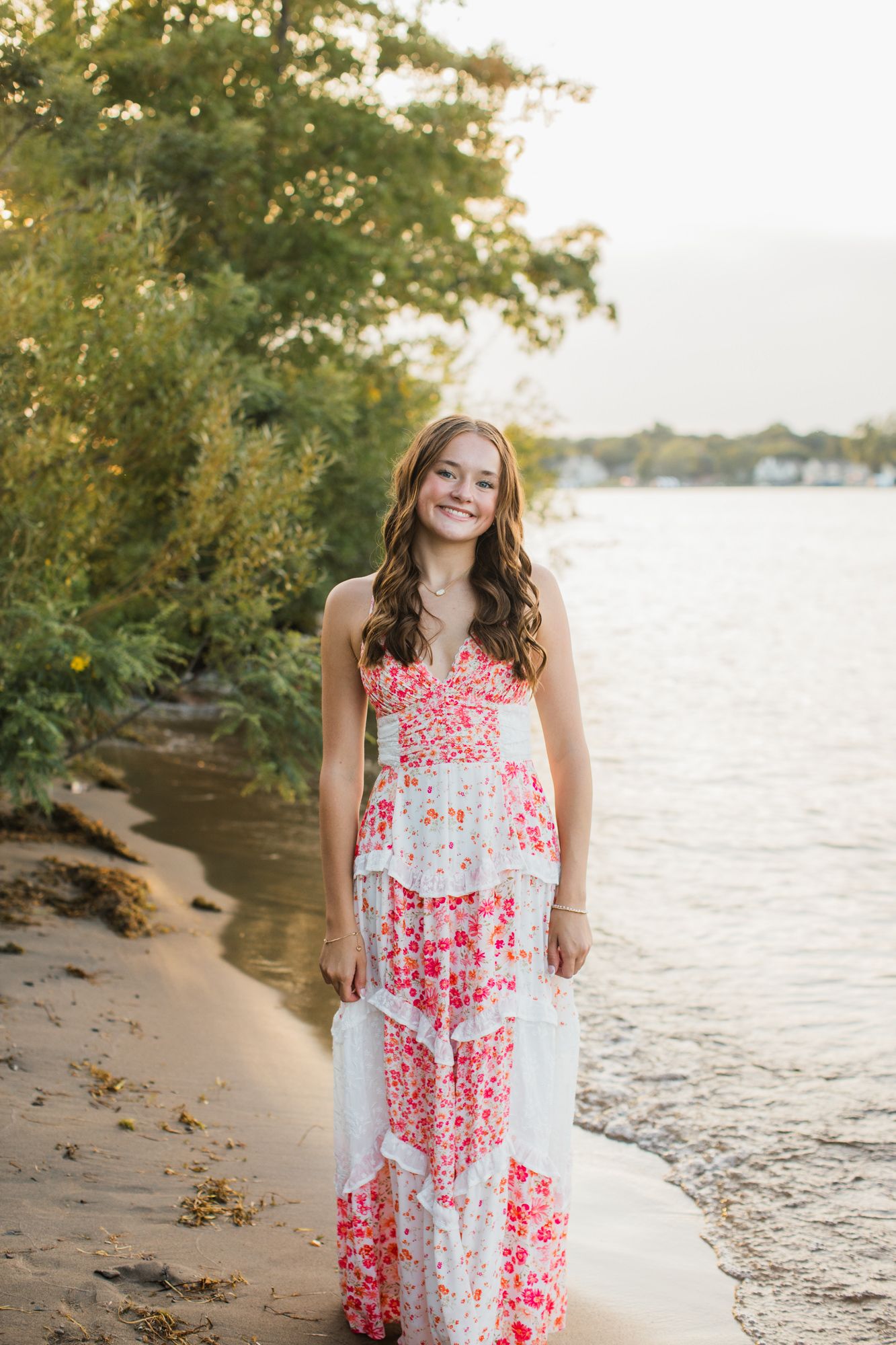 Young woman in a floral maxi dress smiling on a sandy beach shoreline at golden hour.