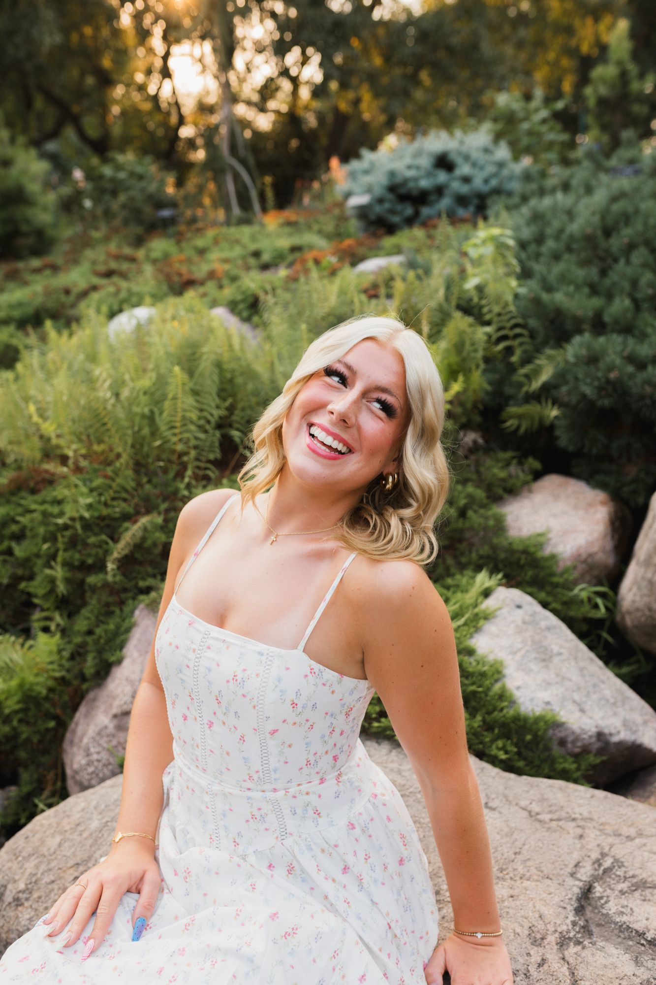 Smiling blonde woman in a floral sundress sitting on rocks surrounded by lush green garden foliage at sunset.