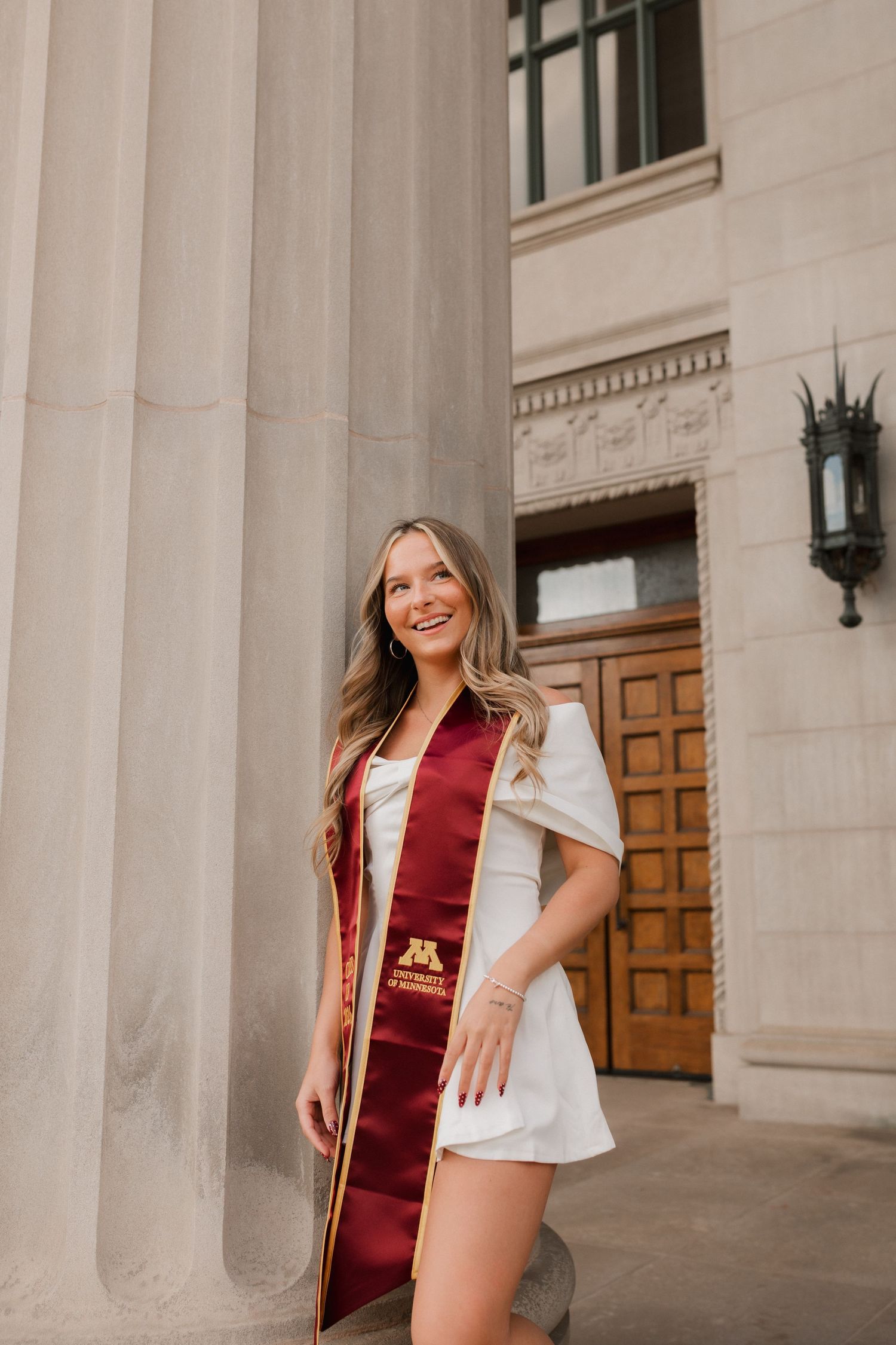 Smiling female graduate wearing a maroon University of Minnesota sash and white dress posing by large stone columns.