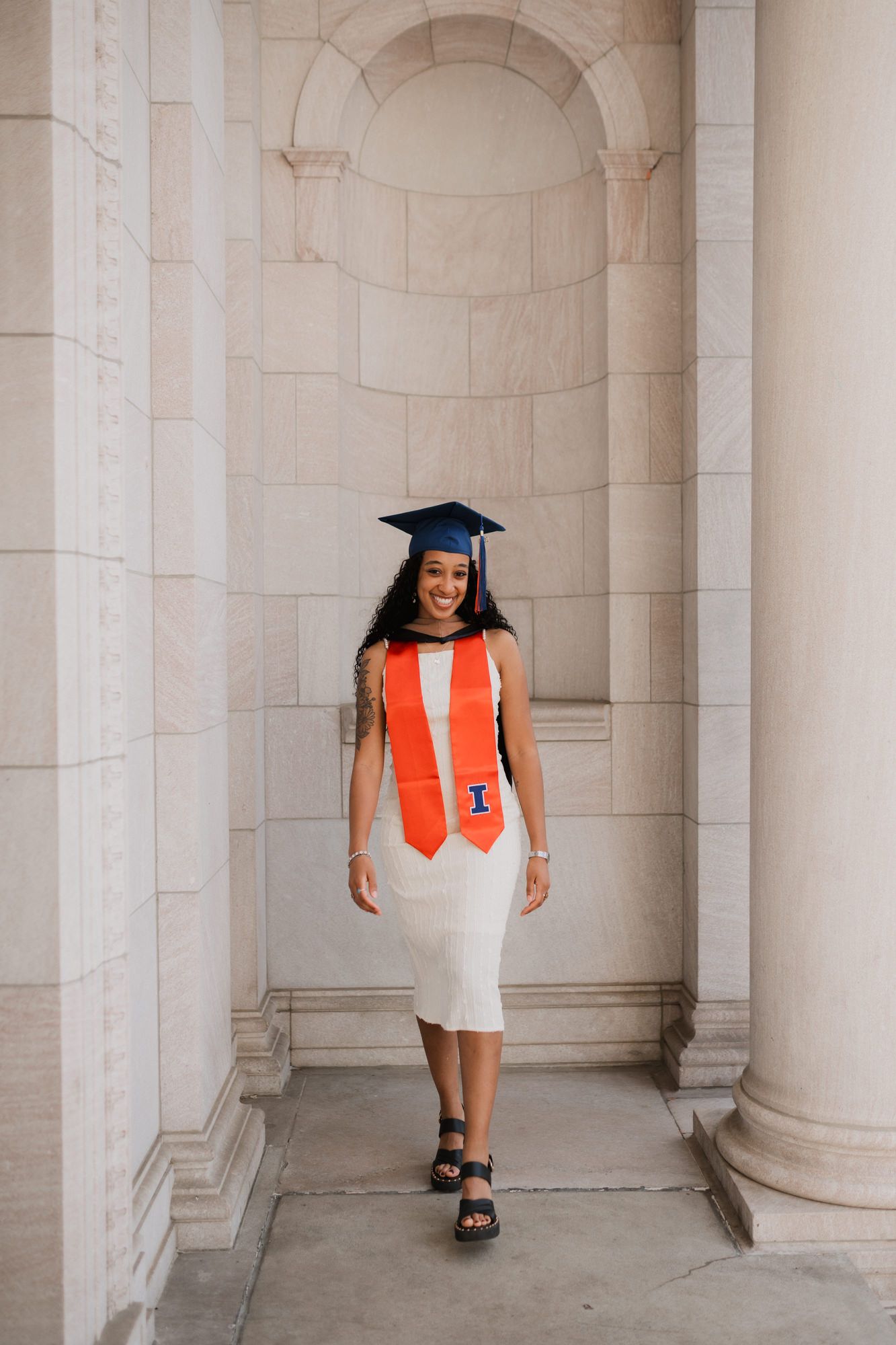 Smiling graduate in white dress, orange University of Illinois stole and cap, posing between stone columns.