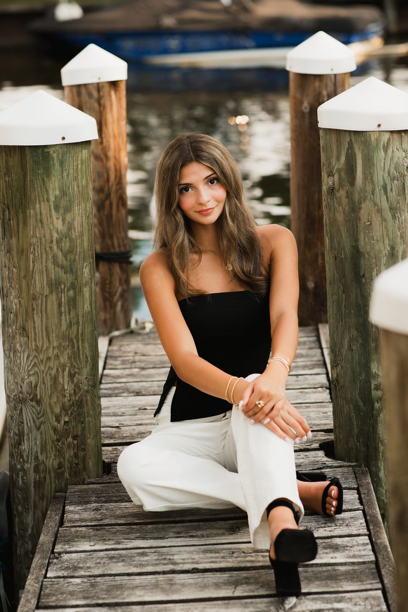 Young woman in black top and white pants posing on a wooden dock near marina pilings.
