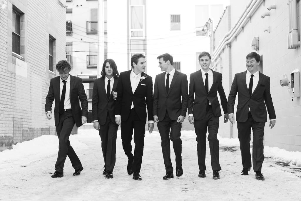 Black and white photo of groomsmen walking in line along urban buildings.