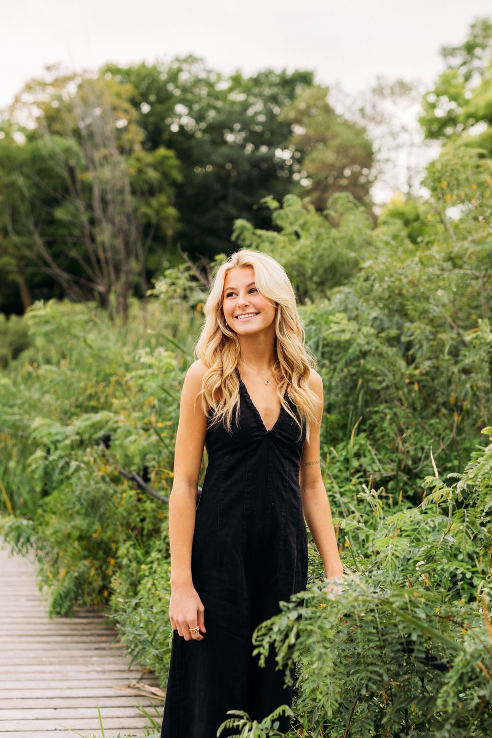 Person in black dress poses among lush greenery on wooden boardwalk with natural lighting.