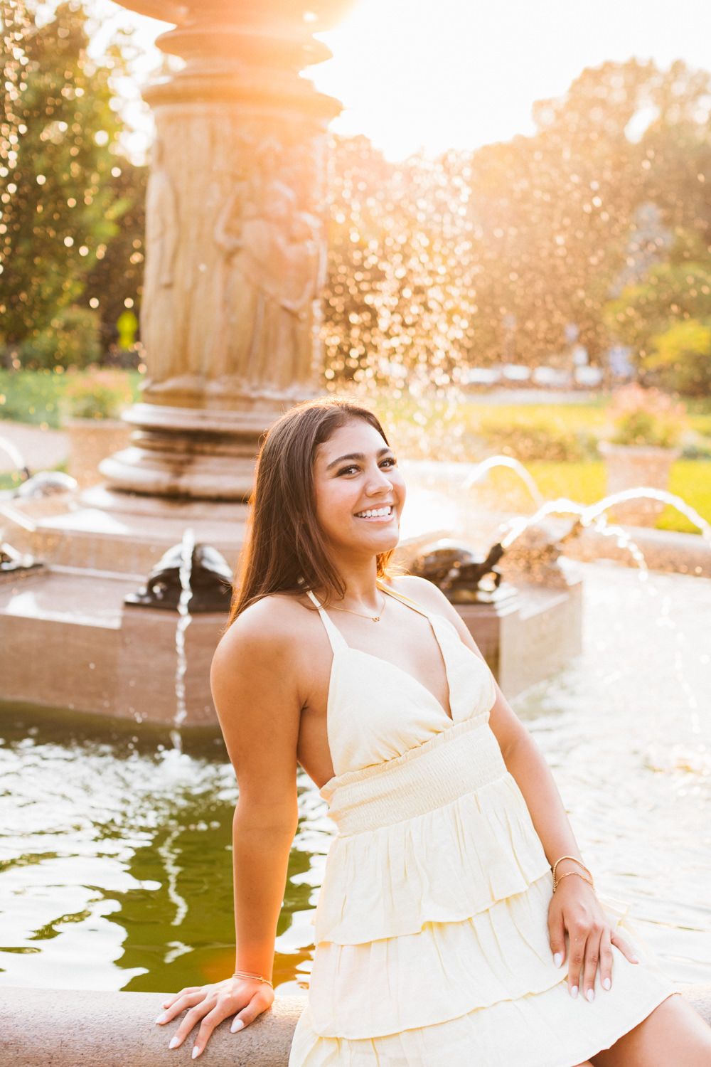 White dress and warm sunlight create dreamy summer scene by ornate fountain in park during golden hour.