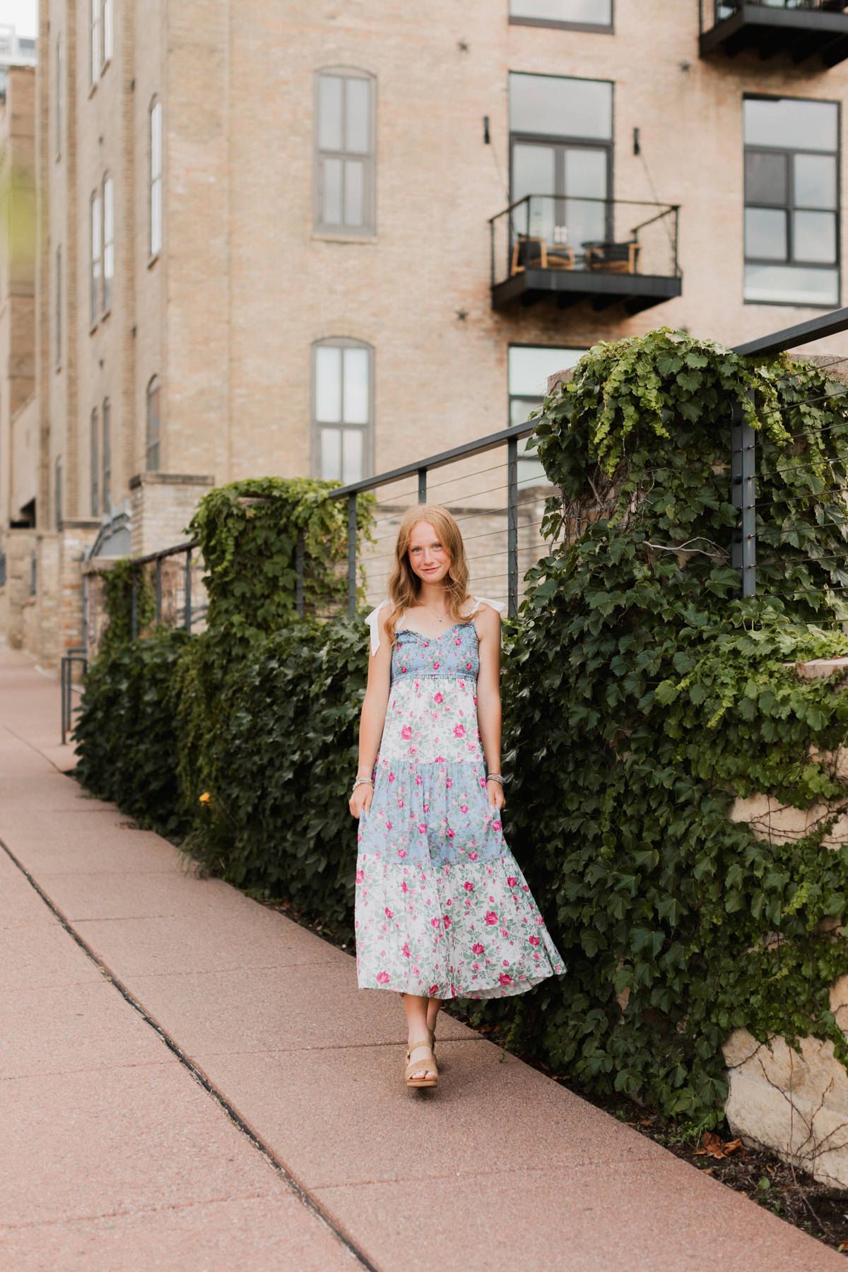 Young woman in blue floral midi dress walking along ivy-covered brick building on urban sidewalk.