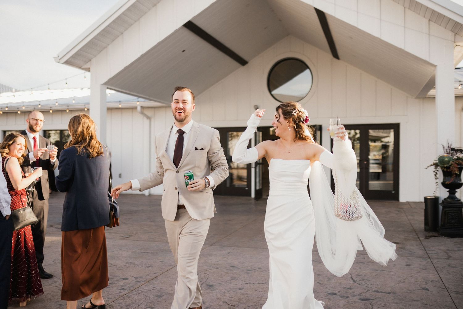 Newlyweds walk hand in hand toward guests outside a white barn venue, the bride raising her arm in joyful celebration.