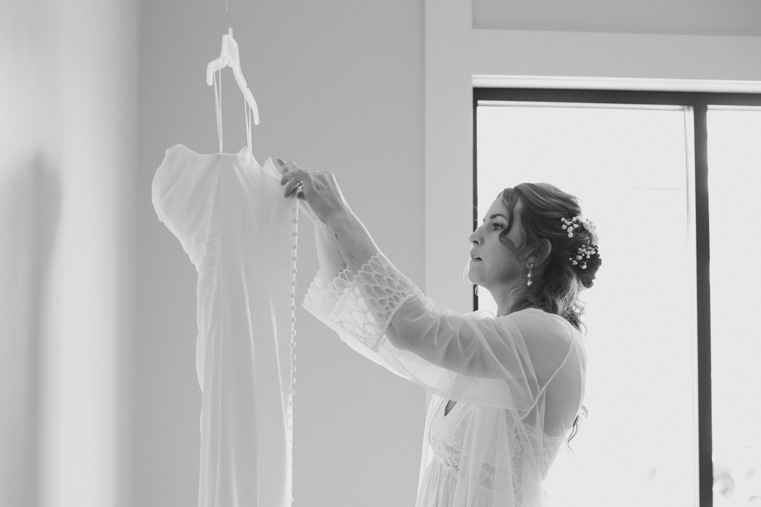 Bride in a lace robe with an updo and floral hair accessory gazes at her wedding dress hanging by a bright window in black and white.