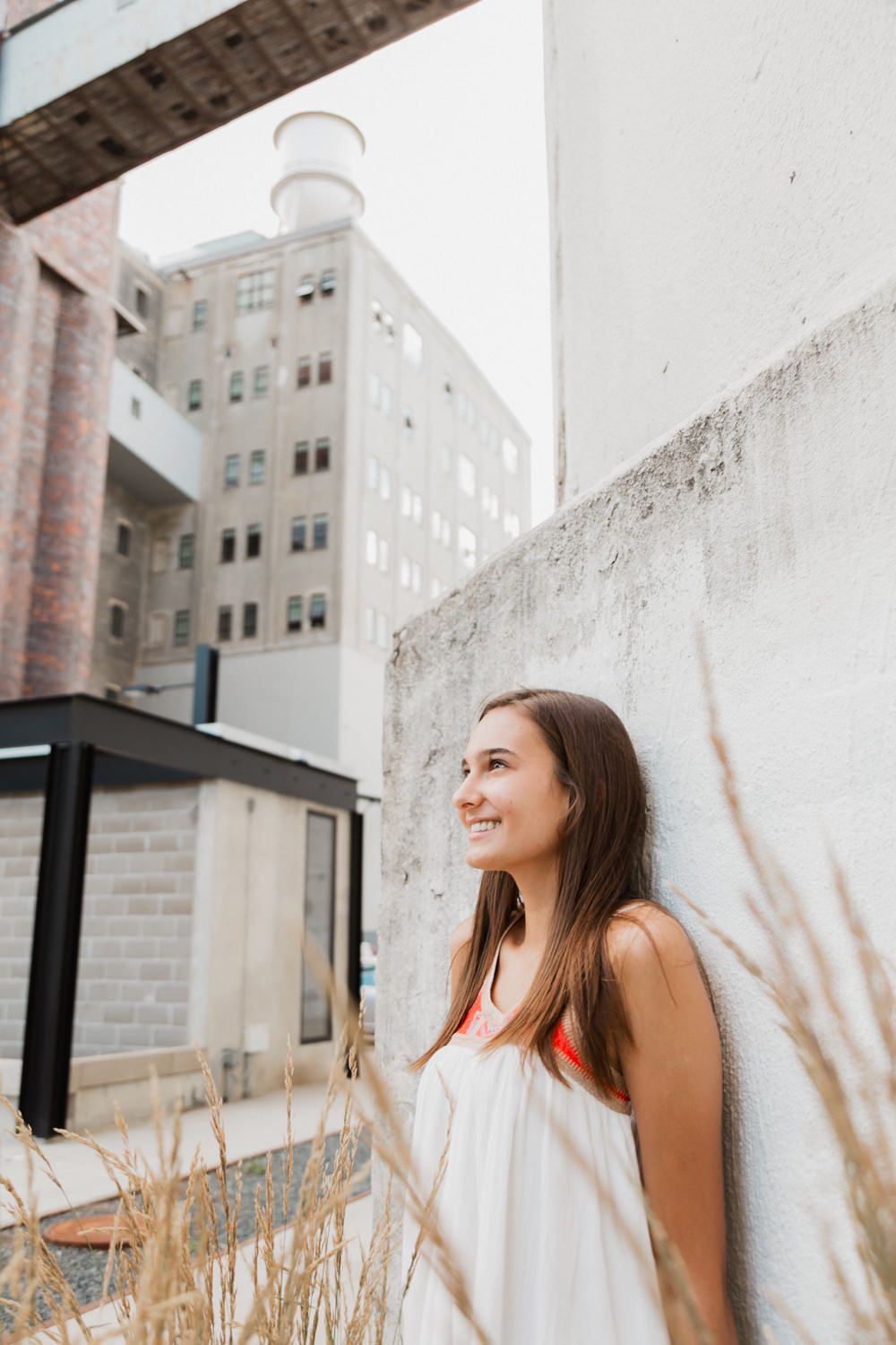 A woman in a white top sits against a concrete wall with tall wheat grass swaying in the foreground.