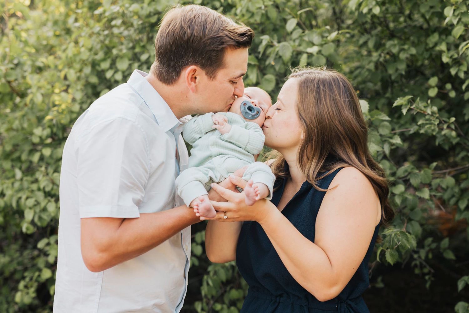 Parents sharing a tender moment with their baby outdoors against a leafy background.