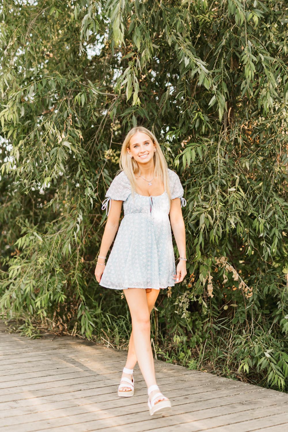 Woman in summer dress walking on wooden boardwalk surrounded by green foliage.