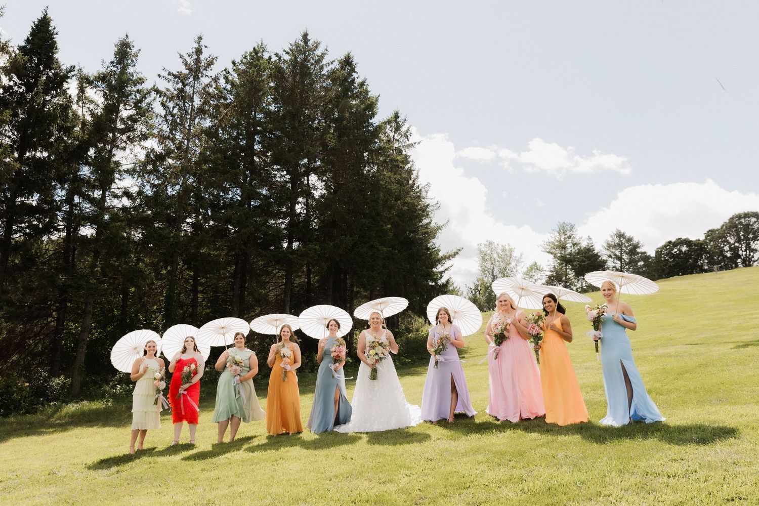 Bridal party in rainbow-colored gowns holding white parasols on a green lawn with tall trees in the background.