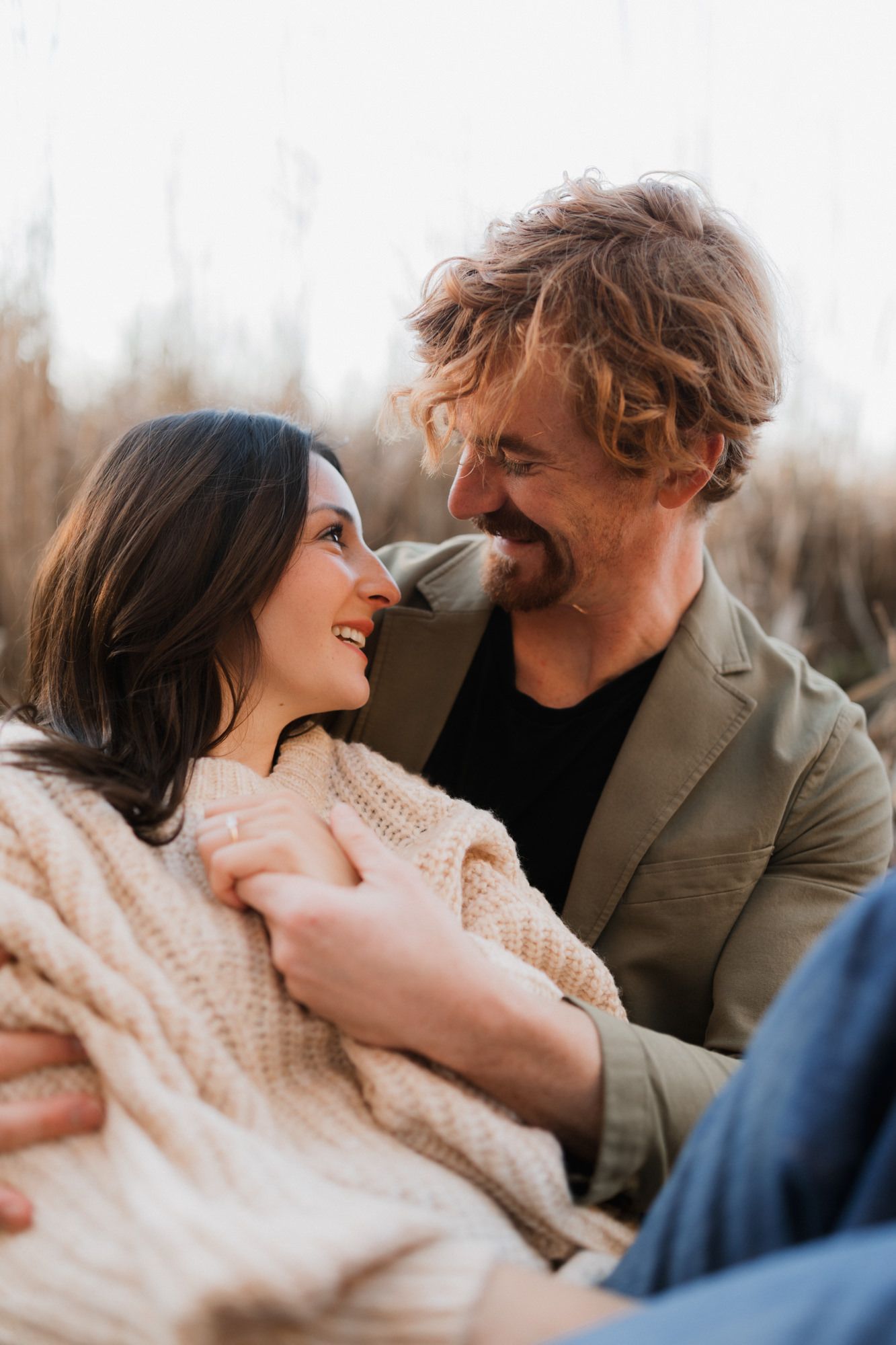 A smiling couple gazes at each other outdoors, woman in cream knit sweater, man in olive jacket, sharing a romantic moment.