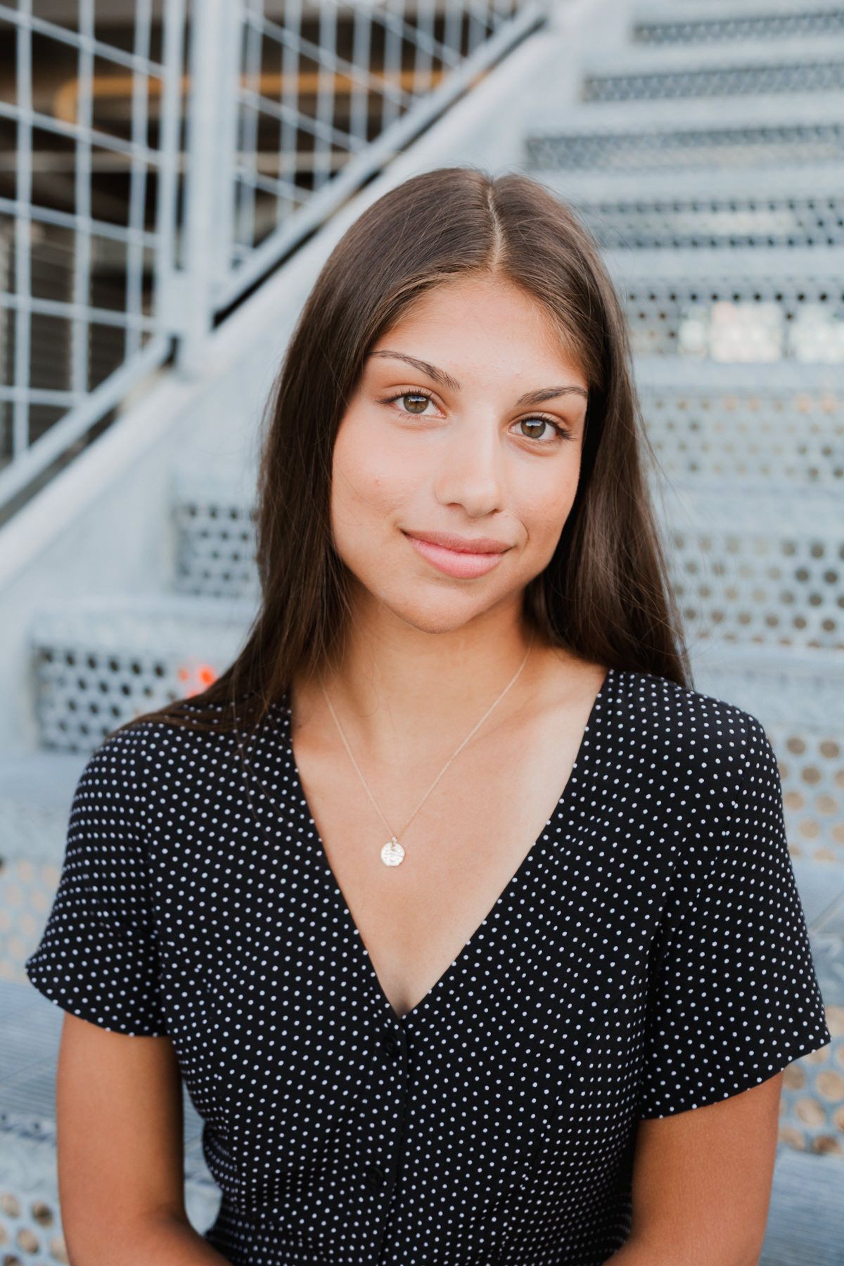Young woman with long dark hair wearing a black polka dot dress and gold necklace on stairs.