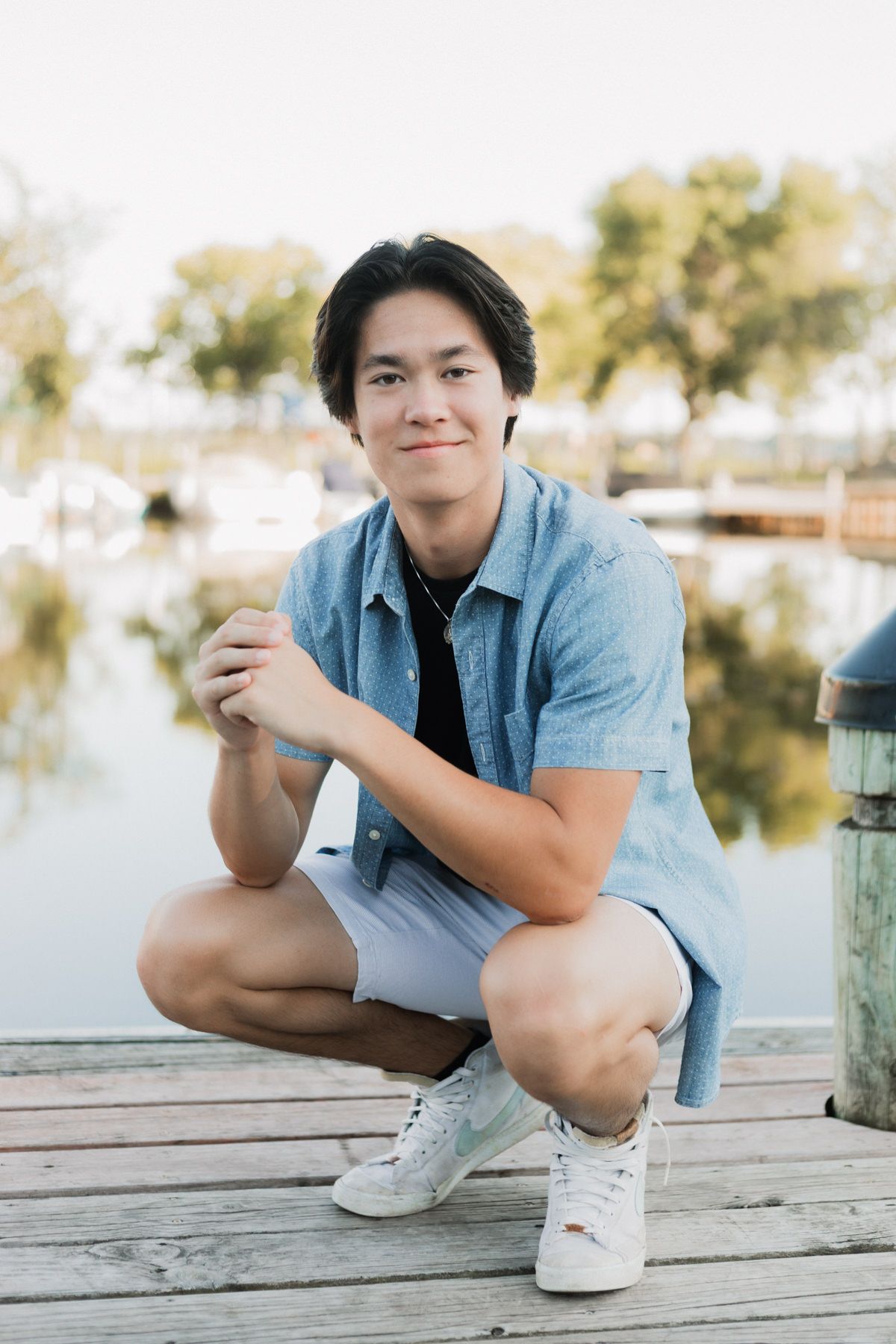 Young Asian man in blue denim shirt and shorts crouching on a wooden dock by a marina, smiling at camera.