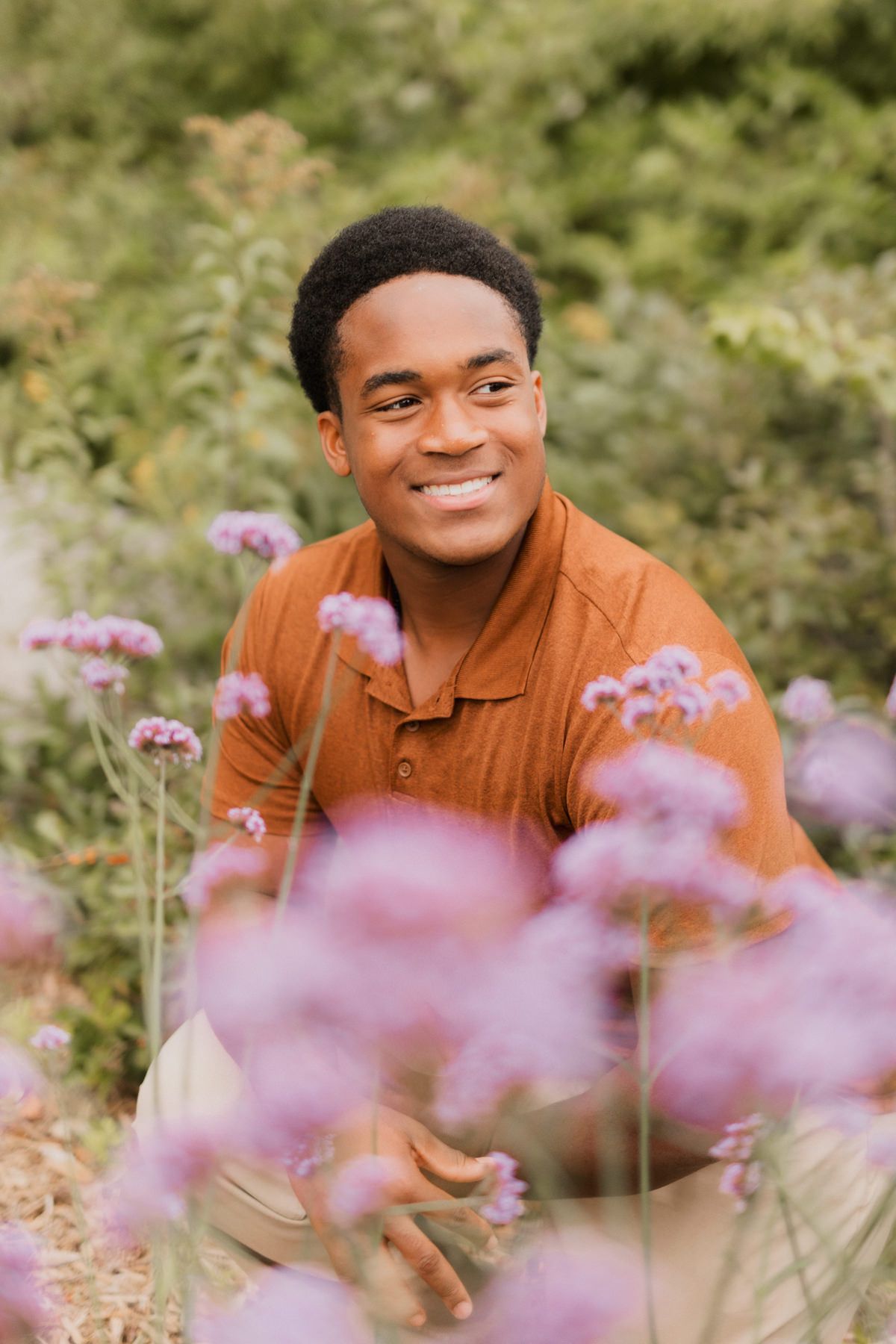 Smiling young man in rust shirt sitting among purple wildflowers in a garden.