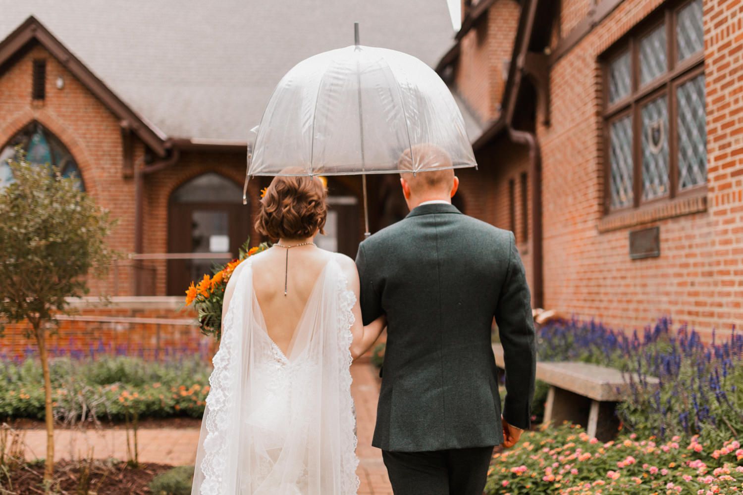 A couple in wedding attire walks through a garden path with brick buildings and autumn foliage in the background.