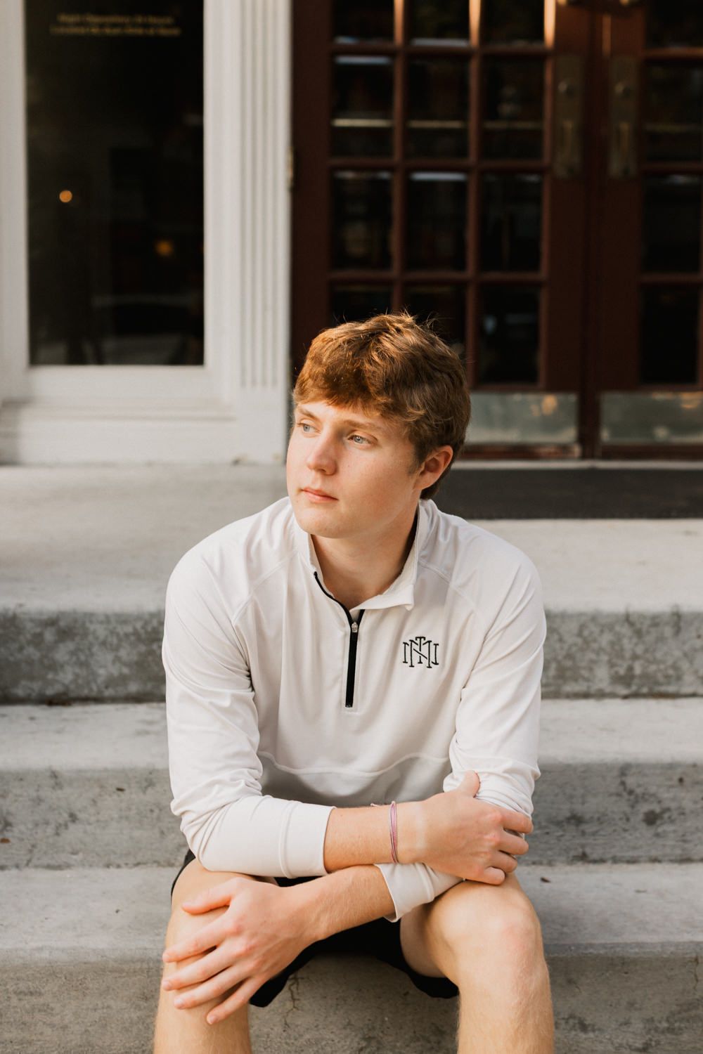 Person in white quarter-zip shirt sitting thoughtfully on steps with windows and brick architecture visible.