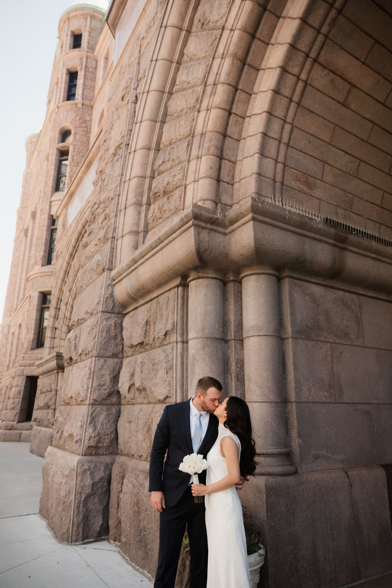 Bride and groom share a romantic kiss outside a grand stone building on their wedding day.