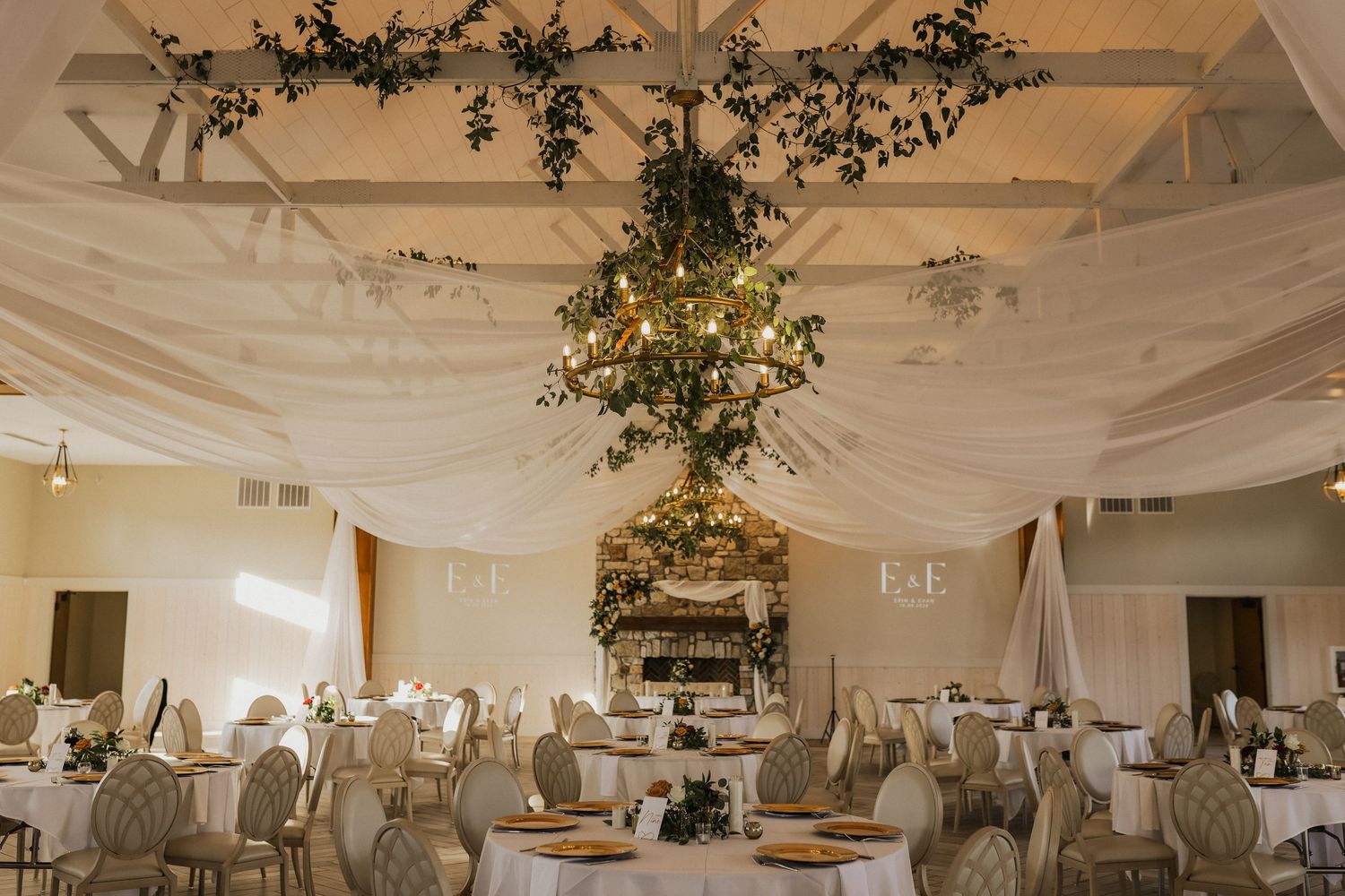 Elegant wedding reception venue with white draped ceiling, hanging greenery chandelier, round tables and chairs set with gold chargers.