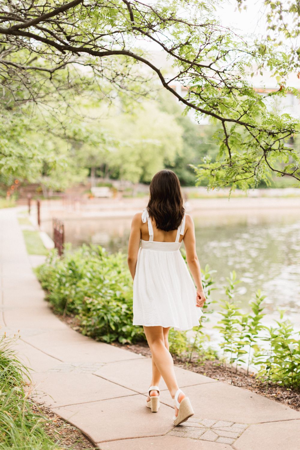 A person in a white sundress walks along a peaceful waterfront path under overhanging tree branches.