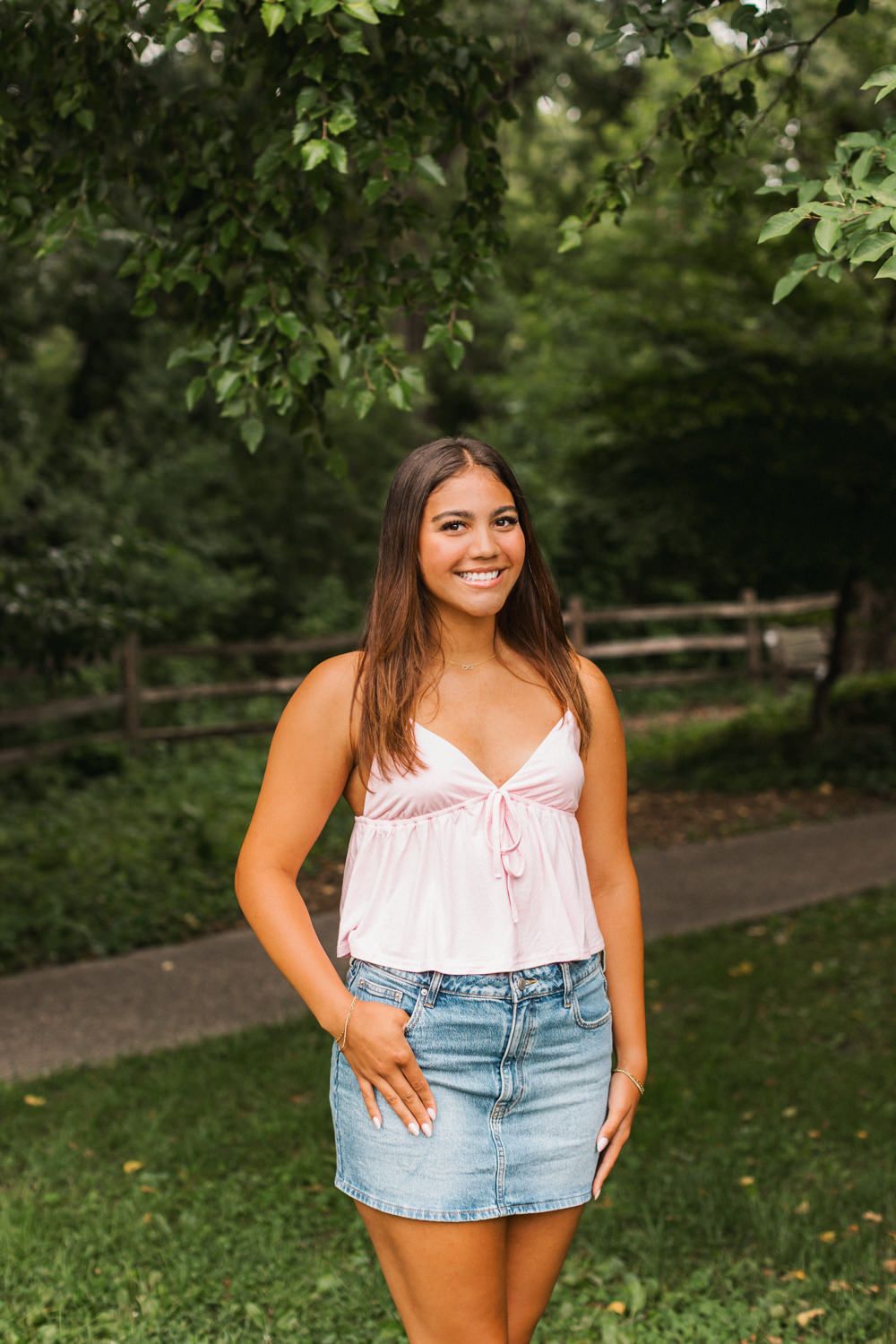 Person in a pink camisole top and denim mini skirt poses outdoors on a tree-lined path during summer.