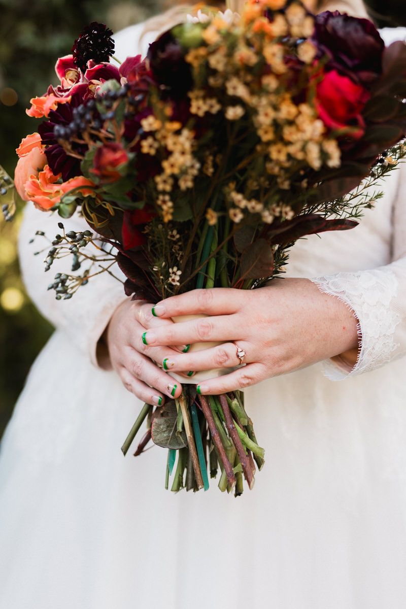 Close-up of bridal bouquet with deep burgundy and orange fall flowers against white wedding dress.