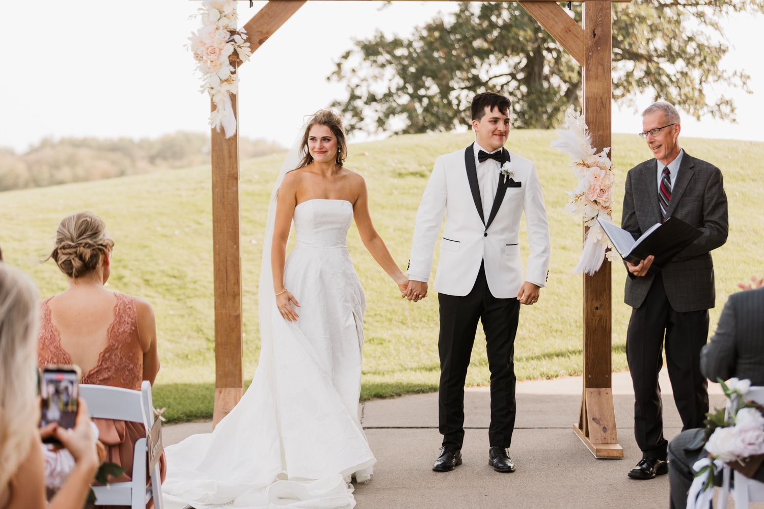 Wedding party in formal attire poses together on a grassy hillside during outdoor ceremony celebration.