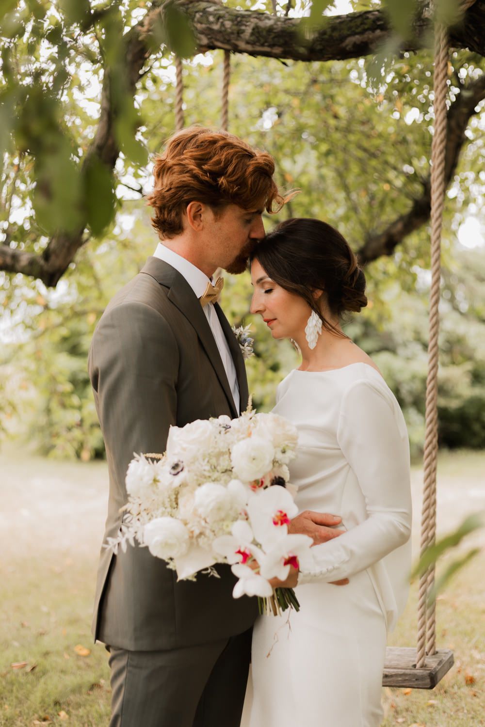 Bride and groom share an intimate moment under a tree swing, bride holding a white floral bouquet outdoors.
