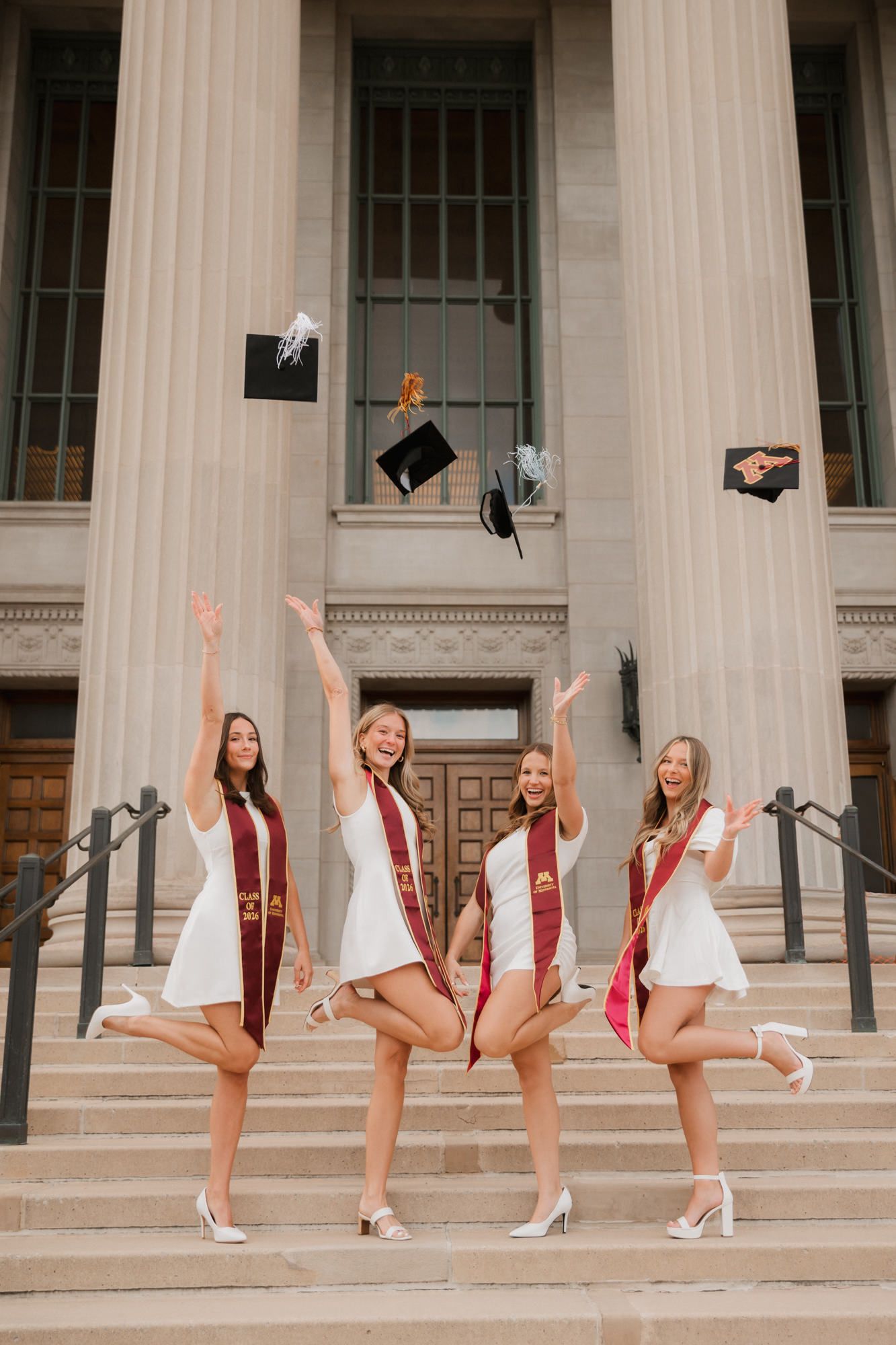 Four happy female graduates tossing caps on university building steps in white dresses and sashes.