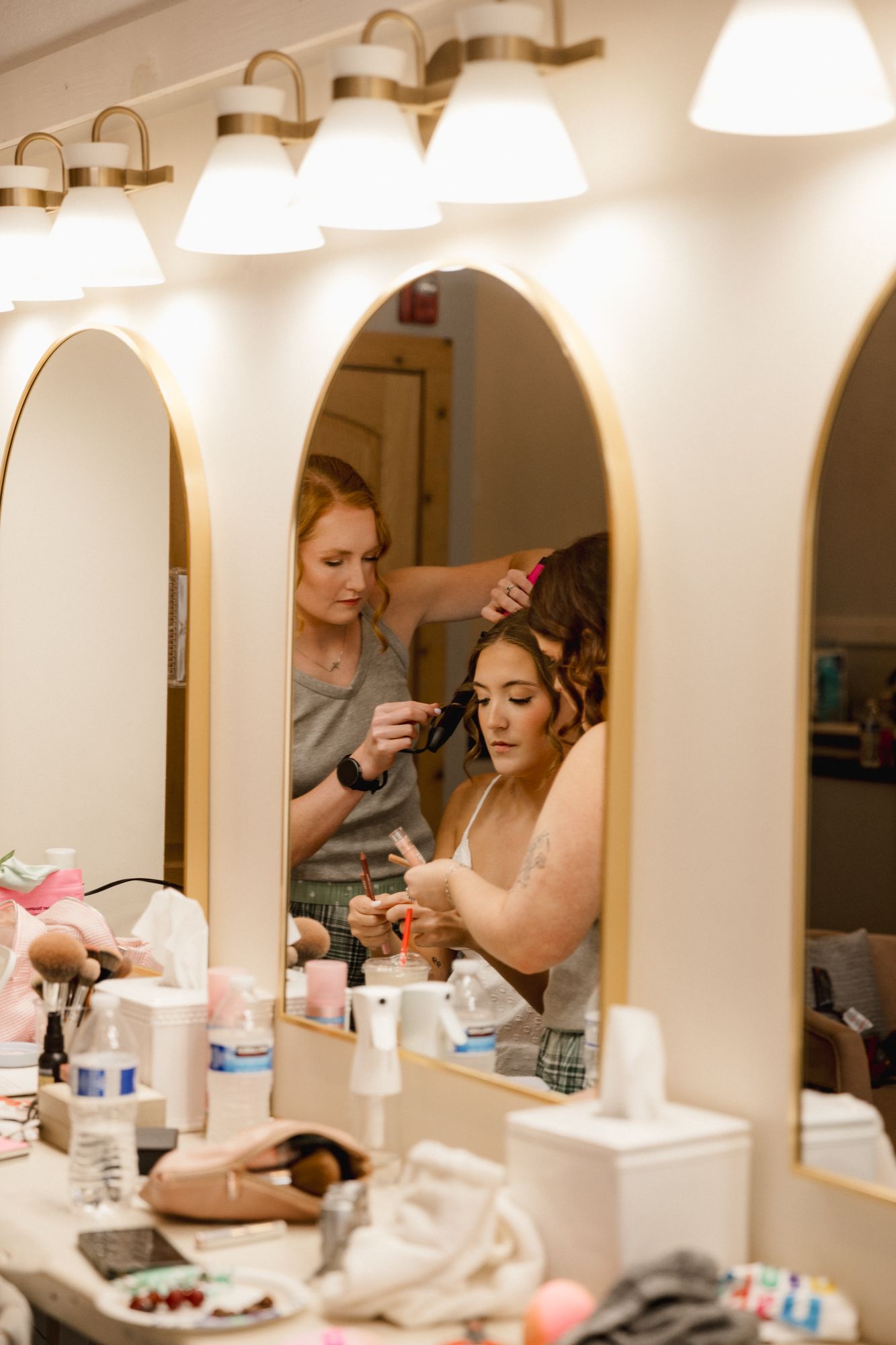 A stylist curls a woman's hair at a vanity mirror with lights, surrounded by beauty products.