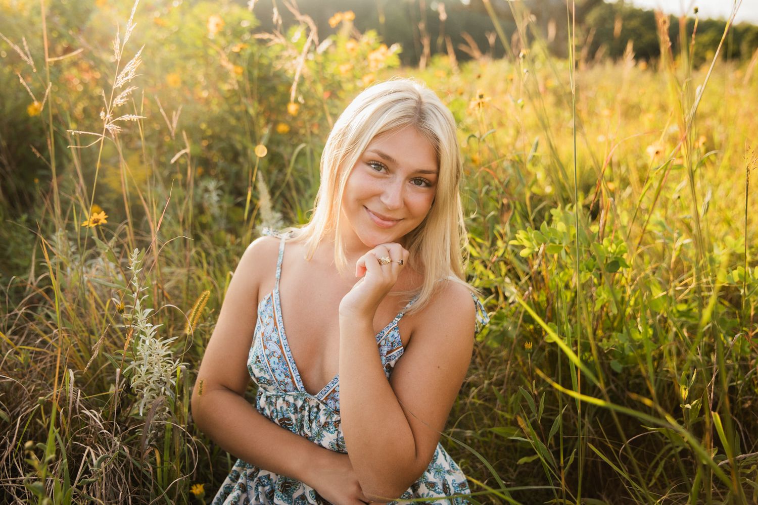 Smiling blonde woman in floral dress sitting in a sunlit meadow with golden wildflowers in the background.