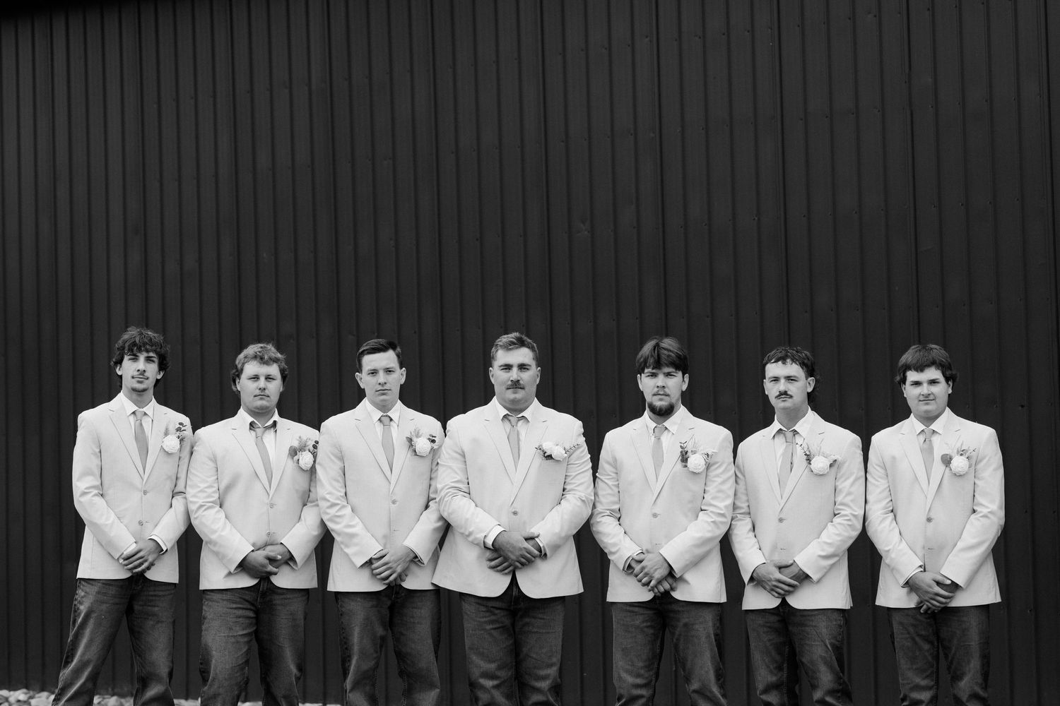 Black and white photo of groom and groomsmen in matching suits with boutonnieres standing in a row outdoors.