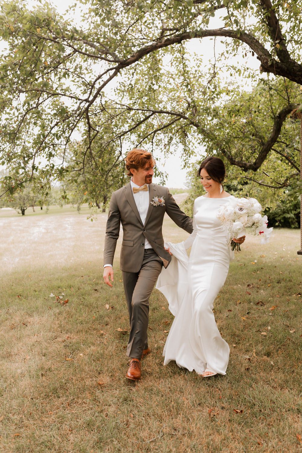 A couple in wedding attire playfully runs and dances together on a grassy hillside under trees.