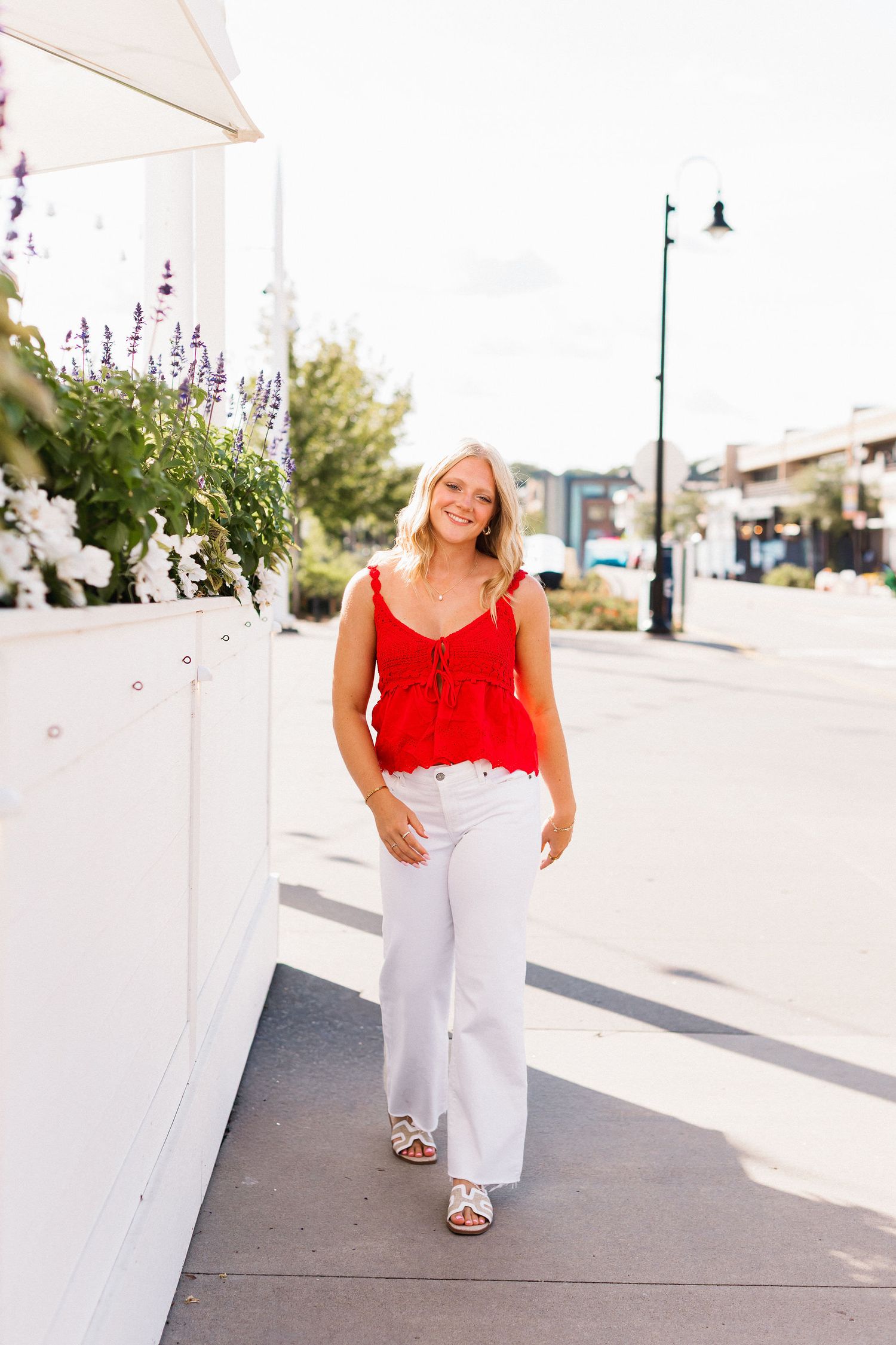 A summer street style outfit featuring a red tank top and white pants paired with sandals on a sunny sidewalk.