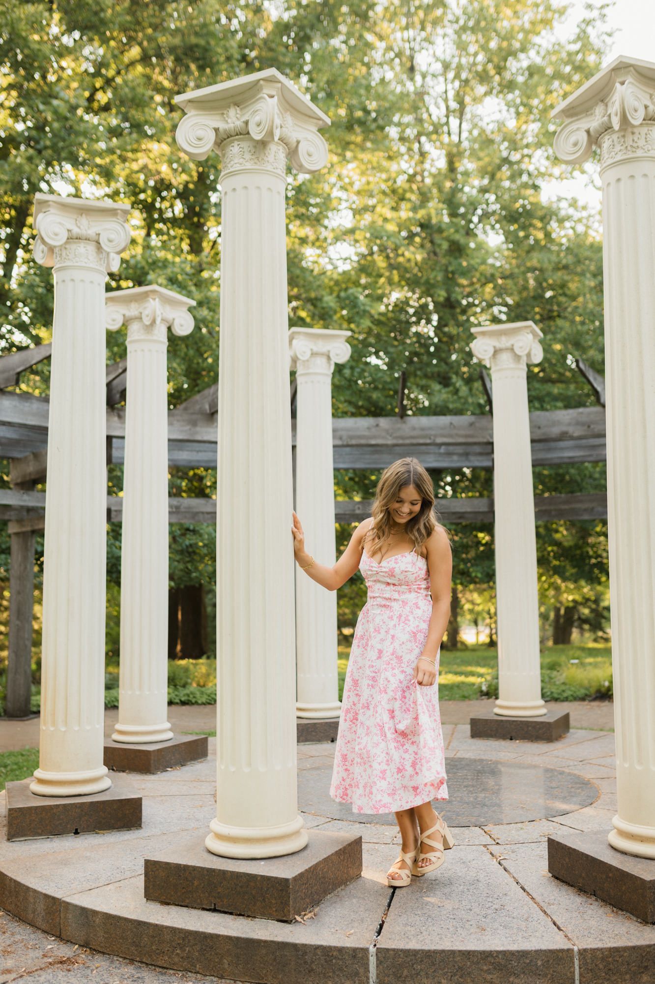 Woman in pink floral midi dress posing among white Ionic columns in a lush green park setting.