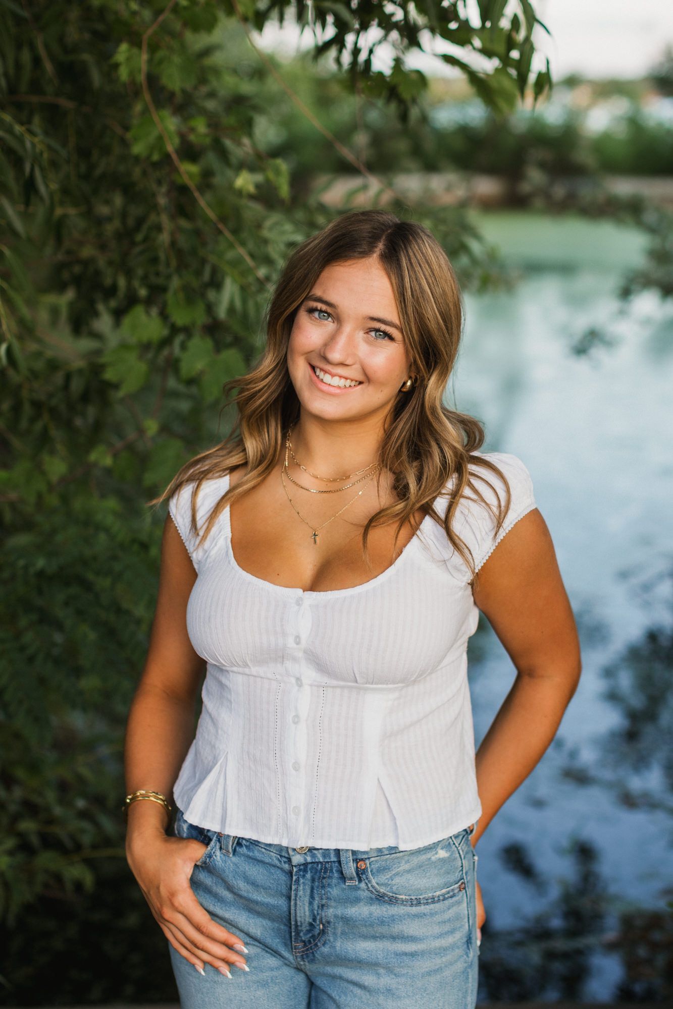 Young woman smiling outdoors in white top and jeans, posing near a pond with lush green trees.