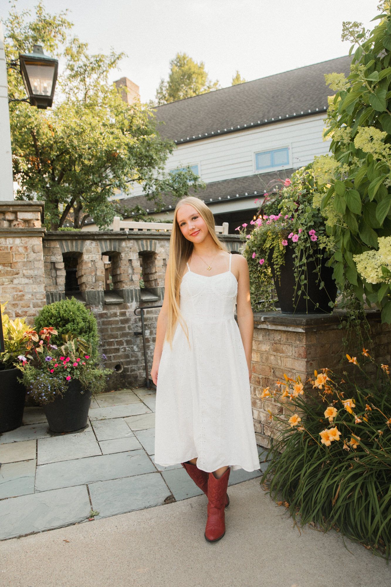 Young blonde girl in white sundress and red cowboy boots posing in a garden with stone walls and flowers.