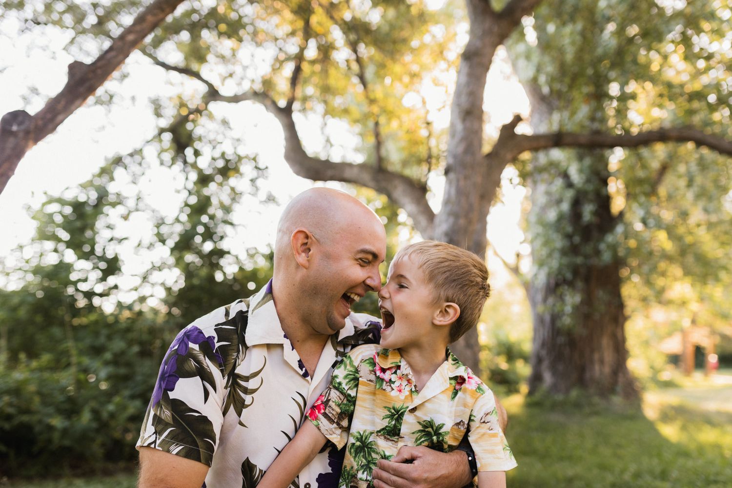Father laughing with young child in matching tropical shirts outdoors in a sunny park.