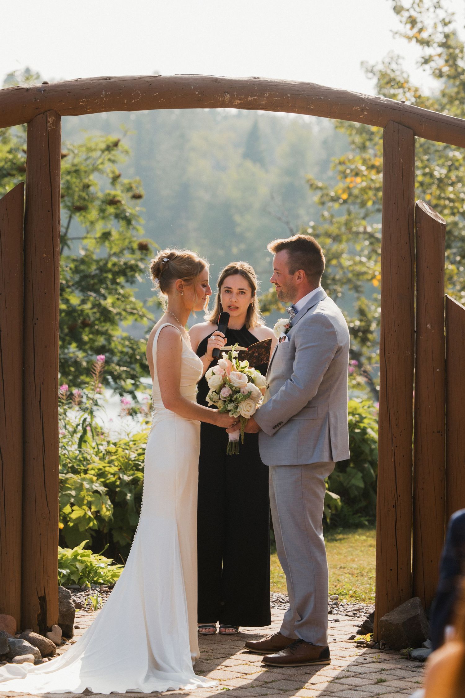 Outdoor wedding ceremony under wooden arch with bride, groom, and officiant surrounded by lush greenery.