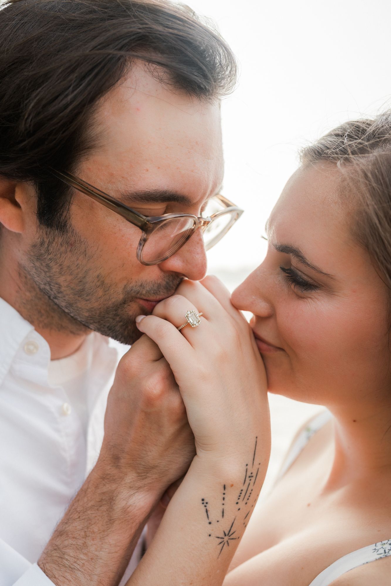 Couple sharing a tender moment, man kissing woman's hand displaying engagement ring, tattoo visible on her arm.
