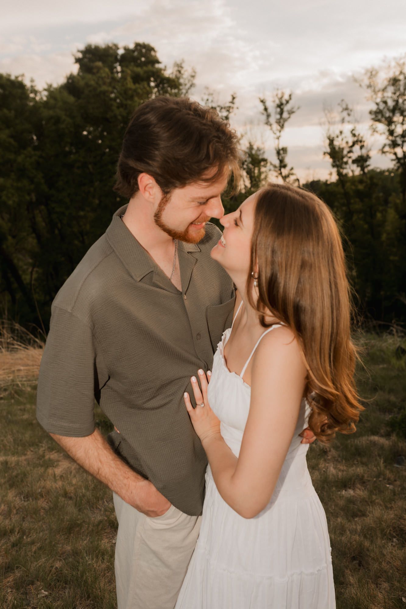 A couple shares a romantic almost-kiss at golden hour in a field, man in olive shirt and woman in white dress.