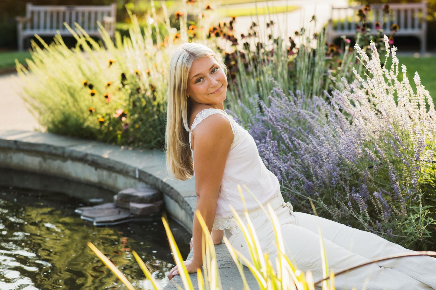 Smiling blonde woman in white dress seated by a garden pond surrounded by lavender and colorful flowers.