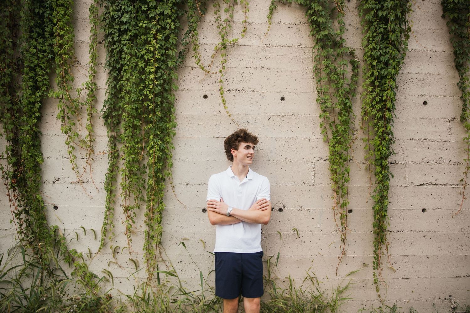 Young man in white shirt and blue shorts stands smiling with arms crossed against ivy-covered concrete wall.