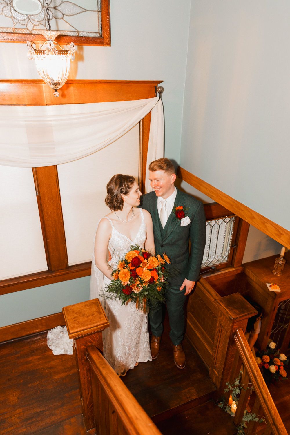 A couple descends a wooden staircase in a historic building with warm wood paneling and blue walls.