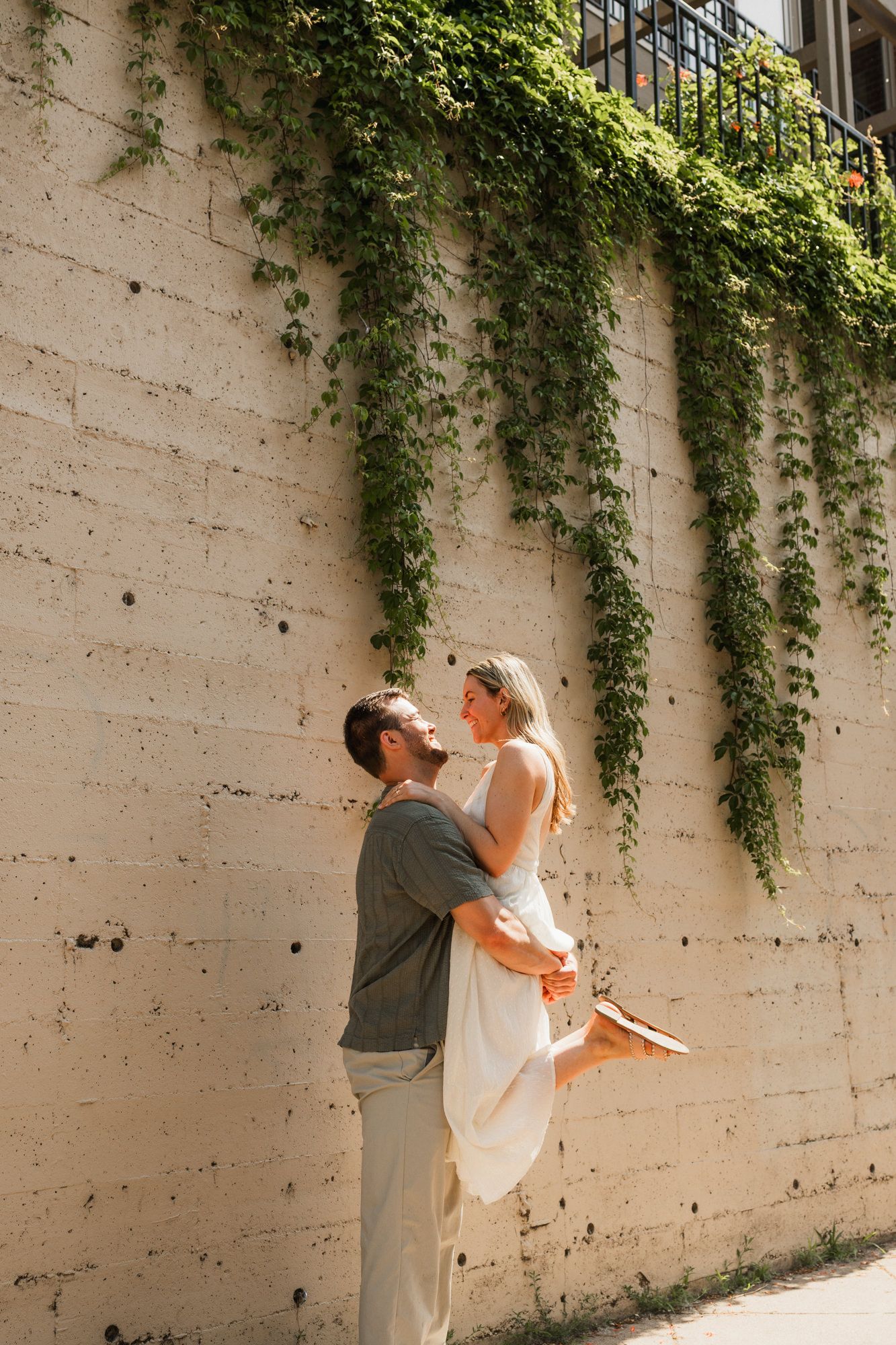 Couple sharing a romantic moment outdoors, man lifting woman in white dress against ivy-covered stone wall.